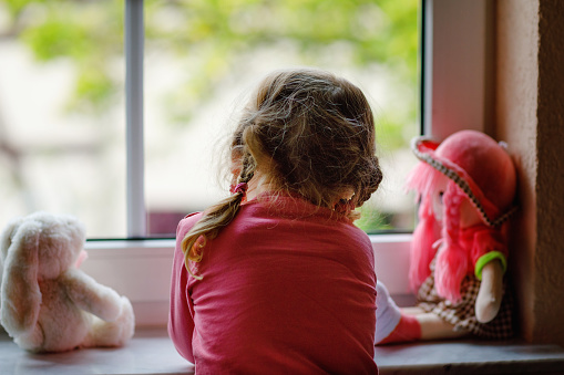 Cute toddler girl sitting by window and looking out on rainy day. Dreaming child with doll and toy feeling lonely. Self isolation concept during corona virus pandemic time. no face, unrecognizable.