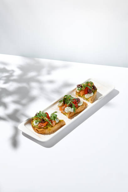 istock Aesthetic composition with salmon bruschetta on white background with shadows from flowers. Italian bruschetta with salmon, avocado, cheese and olives on fine dining in summer. Elegant menu concept. 1418761434