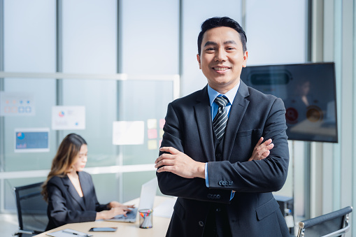 Asian businessman or manager in suit arms crossed smiling standing in modern office with employee or colleague