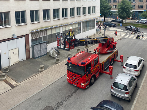 Stockholm, Sweden - August 27, 2022: Aerial view of a firefighter on a red fire engine ladder about to enter a residential building in Stockholm Sweden August 27, 2022. Incidental people in the background.