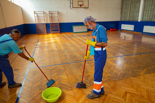 A rear view of two senior male janitors cleaning the wooden floor of a large and empty sports hall with the broom and disinfectant.