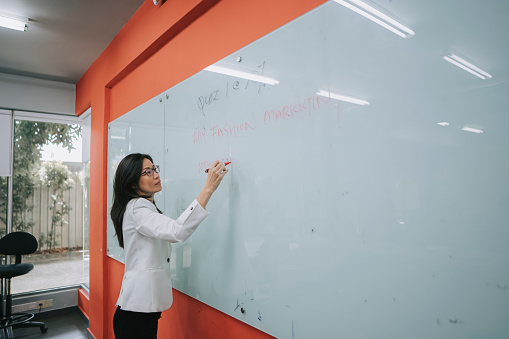 Asian Chinese female Lecturer writing on whiteboard in classroom explaining to her student