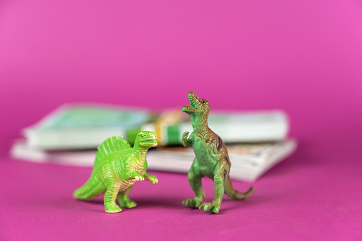 Dinosaur miniatures and wads of bills against a purple background. Wads of cash in the background . Green figures of animals of prey standing on their hind legs. Selective focus.