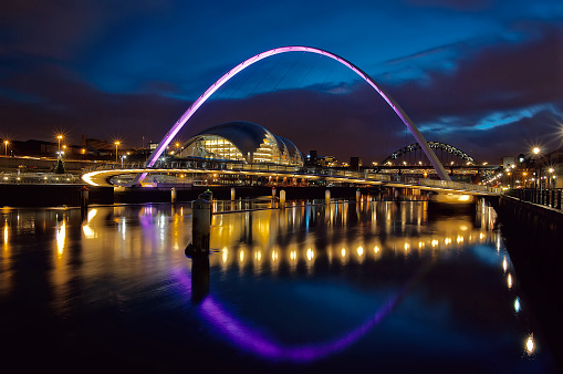 The Gateshead Millennium Bridge at blue hour, low light, displaying pink neon light reflected in the river tyne - long exposure, a pedestrian and cyclist tilt bridge spanning the River Tyne between Gateshead arts quarter on the south bank and Newcastle upon Tyne's Quayside area on the north bank. It was the first tilting bridge ever to be constructed