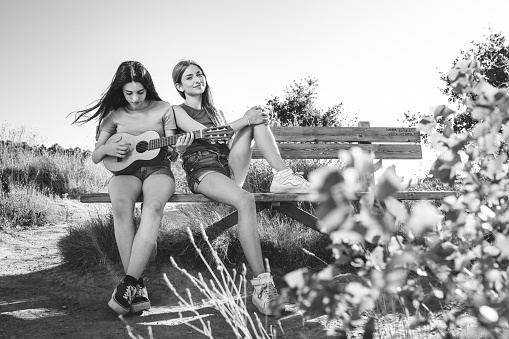 Young beautiful girl playing guitar and other looking her, sitting on a wooden bench outdoors. Friendship concept. Monochrome version.