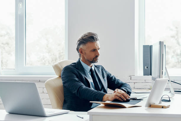 istock Confident mature man in formal business wear using computer while sitting at his working place 1418804748