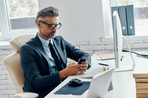 istock Confident mature man in formalwear using smart phone while sitting in office 1418804849