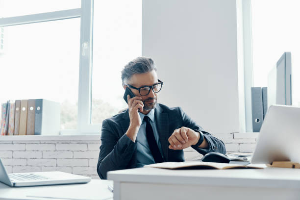 istock Confident mature man in formalwear checking the time while talking on mobile phone in the office 1418804901