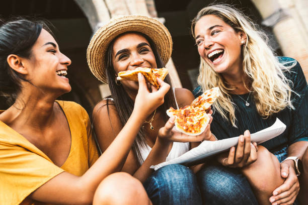 Happy friends eating street food on summer vacation - Three women eating pizza slice on city street - Happy lifestyle and tourism concept stock photo