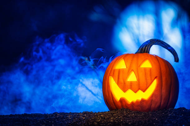 A glowing smiling pumpkin Jack O’ Lantern sitting in a field with a moon lit low fog and the glow of a rising full moon in the background.