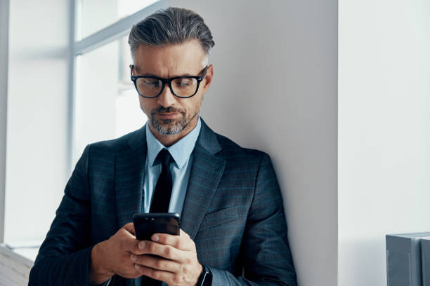 istock Confident mature man in formalwear using smart phone while standing near the window in office 1418807314