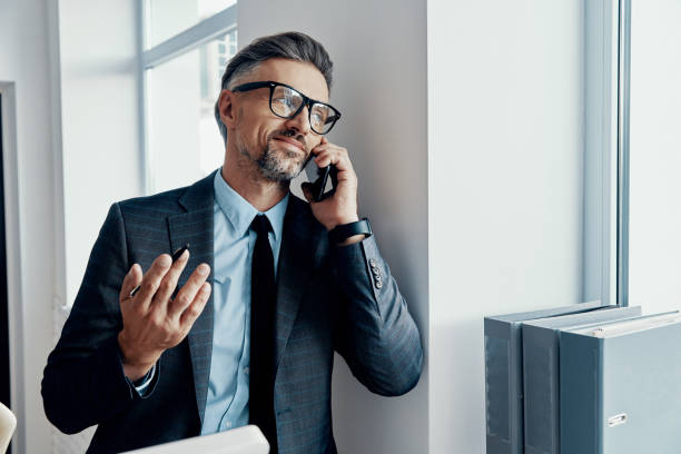 istock Cheerful businessman talking on mobile phone and gesturing while standing near the window 1418807361