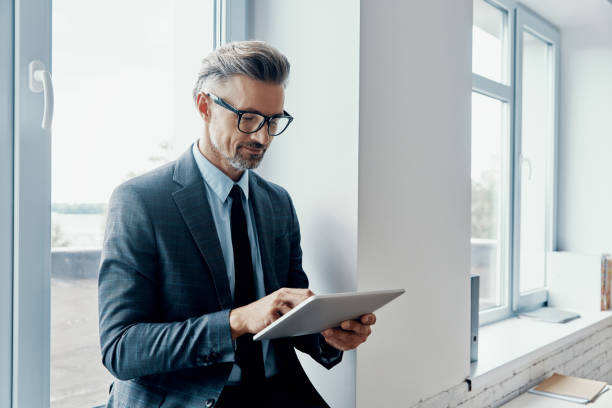Concentrated mature man in formalwear using digital tablet while standing near the window in office stock photo