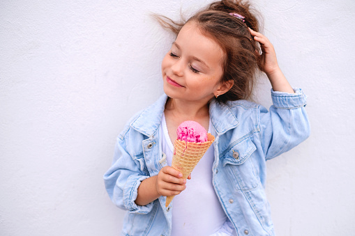 A girl in a denim blue jacket with a horn and a ball of pink ice cream straightens her hair flying in the wind, standing against a white concrete wall. Enjoying the milky creamy taste