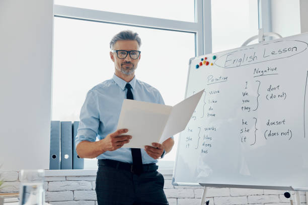 istock Confident English teacher holding papers while standing near the whiteboard at classroom 1418808900