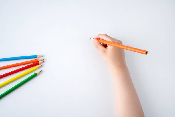 Close up photo of unrecognizable young girl sitting at desk and drawing something with colored pencils on a white paper at home