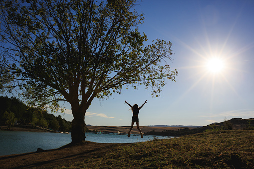 Silhouette of a girl girl jumping on the lake shore under a tree with a starry sun. A girl stands on the shore.