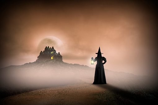 A witch holds a lantern to guide her way on the dirt road to her haunted house high on the hill in the distance.