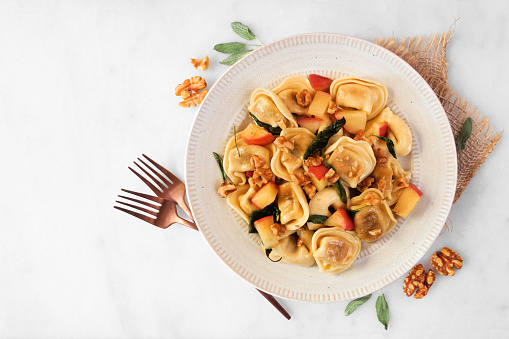 Fall pumpkin and apple tortellini pasta with walnuts and brown butter sage sauce. Top view with frame of ingredients on a white marble background.