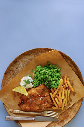 Stock photo showing close-up, elevated view of a wooden platter lined with greaseproof paper that is filled with a portion of battered cod and chips, with a lemon slice, mushy peas and tartare sauce, against a blue background.