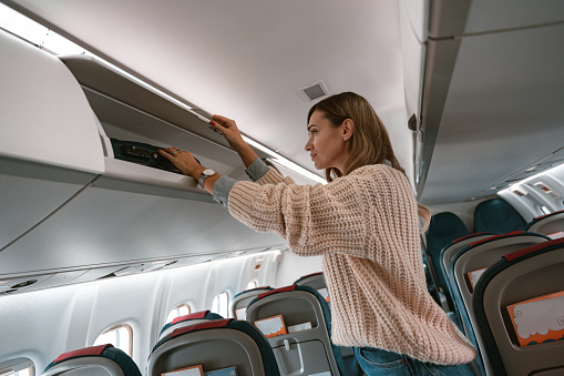 Woman traveler putting luggage into overhead locker on airplane during boarding. High quality photo