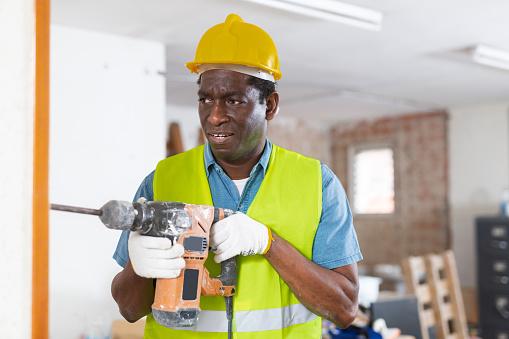 Confused inexperienced african american builder working in building under renovation, trying to remove concrete layer from wall with pneumatic demolition hammer