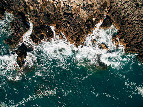 Small waves repeatedly crash on a small sandy beach in a bay with an uneven rocky coastline, aerial photography overhead. High quality photo