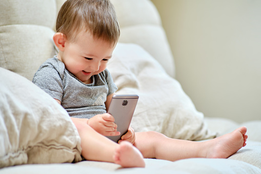 Happy toddler baby boy is sitting with a phone on the sofa in the living room. Child with a smartphone in his hands on the bed