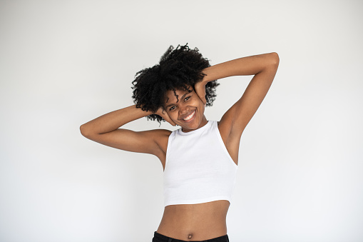 Portrait of happy young African American woman smiling at camera. Female model wearing white crop top and jeans standing with hands behind head against white background. Happiness concept