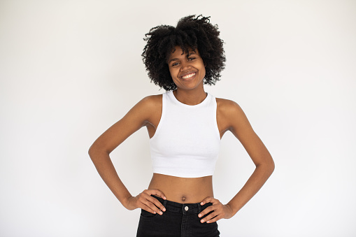Portrait of positive young African American woman smiling. Female model wearing white crop top and jeans standing against white background. Happiness concept