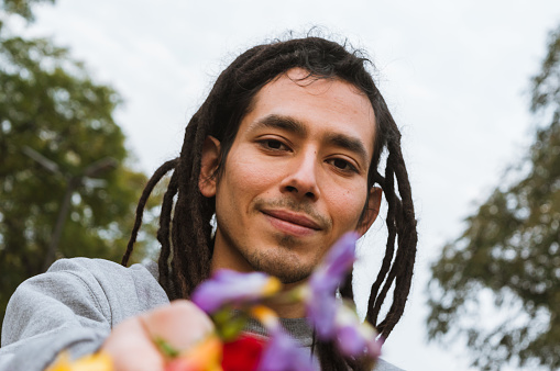 portrait of young venezuelan man with dreadlocks smiling and looking at the camera, outdoors holding flowers with his hand, concept of diversity and lifestyles.