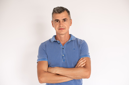 Portrait of confident mature man standing with folded arms. Caucasian man wearing blue T-shirt standing and looking at camera over white background. Confidence concept