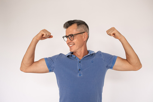 Portrait of smiling mature man wearing glasses showing muscles. Caucasian man wearing blue T-shirt showing biceps and looking away. Power concept