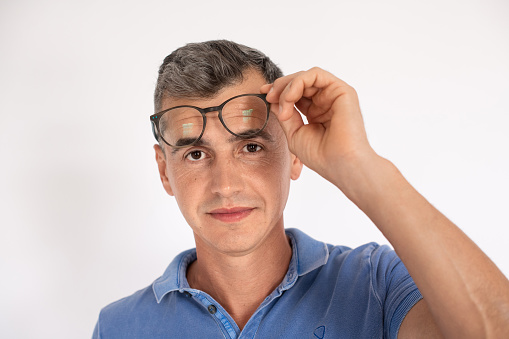 Portrait of mature man taking off eyeglasses looking at camera. Caucasian man wearing blue T-shirt standing over white background. Vision correction concept