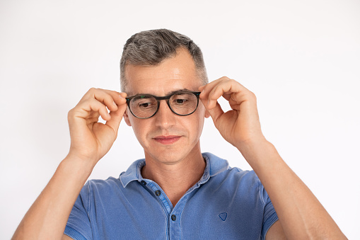 Portrait of mature man wearing blue T-shirt putting on glasses. Caucasian man wearing eyeglasses standing over white background. Eyesight concept