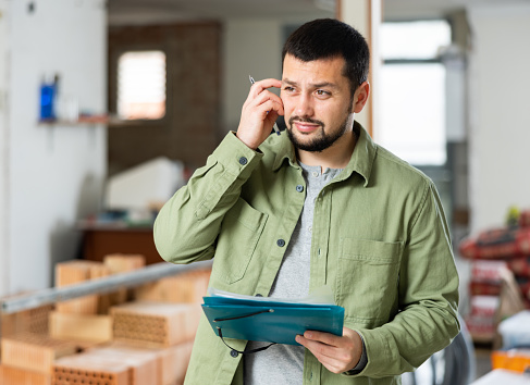 Puzzled and confused young bearded man standing in his house with papers in hands during renovations, determining scope of work