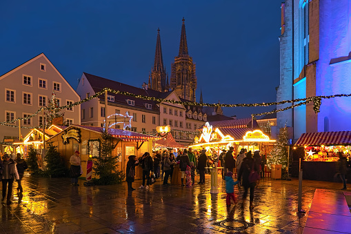 Regensburg, Germany - December 13, 2019: Christmas market at Neupfarrplatz (New Parish Square) close to Neupfarrkirche (New Parish Church) in dusk. Towers of Regensburg Cathedral are visible in the background. Unknown people walk around the market stalls.