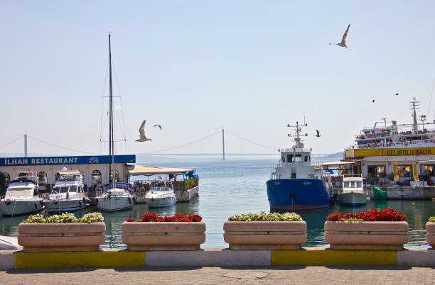 Çanakkale, Turkey - August 18, 2022: Gallipoli Gelibolu center view in a summer day, people near the harbor on the  pavement, there are lots of boat in marina and fish restaurant during the way. You see 1915 Canakkale Bridge from harbor.
