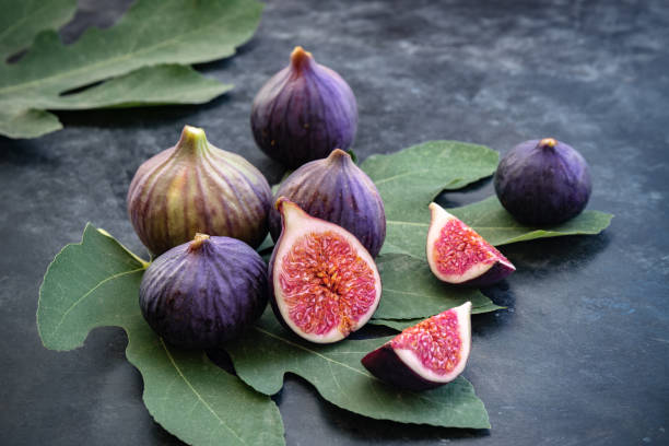 ganze und in scheiben geschnittene feigen auf großen feigenblättern. frische reife früchte auf dunklem hintergrund. nahaufnahme. selektiver fokus. unscharfer hintergrund. - fig leaf stock-fotos und bilder