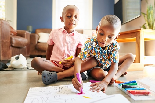 Two cute little African boys colouring together in a drawing book in their living room at home