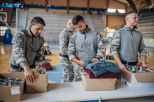 Group of people, soldiers on humanitarian aid preparing donations for civilians in school gymnasium, after natural disaster happened in city.