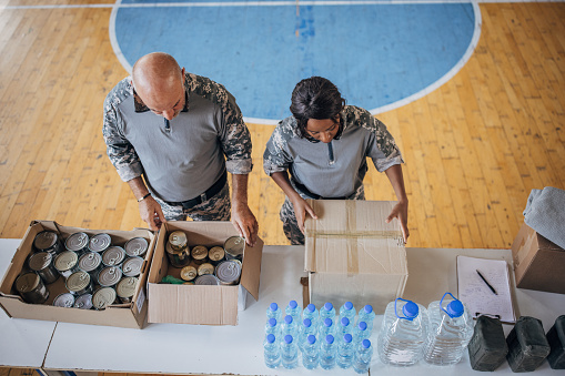 Male and female soldiers on humanitarian aid preparing donations for civilians in school gymnasium, after natural disaster happened in city.