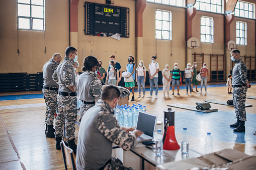Diverse group of people, soldiers on humanitarian aid to civilians in school gymnasium, after natural disaster happened in city.