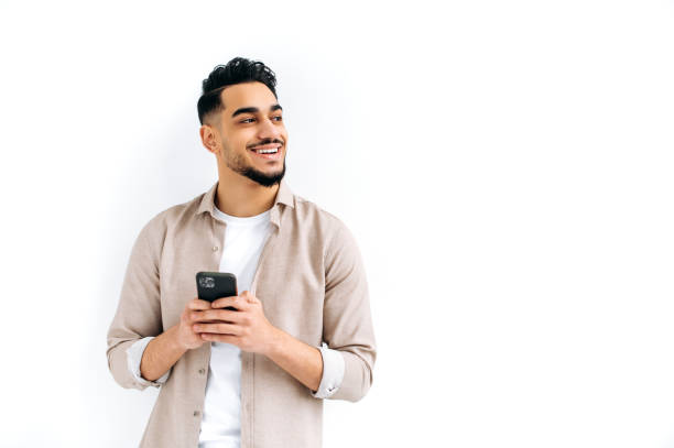 istock Positive attractive indian or arabian guy, holding smartphone in hand, chatting online, browsing internet, looking to the side, thinking, dreaming, standing on isolated white background, smiling 1418863834
