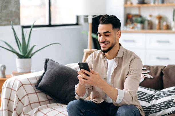 istock Positive attractive friendly indian or arabian guy, stylishly dressed, sitting on a sofa in interior living room, using his smartphone, chatting on social media with friends, browsing online shopping 1418863853
