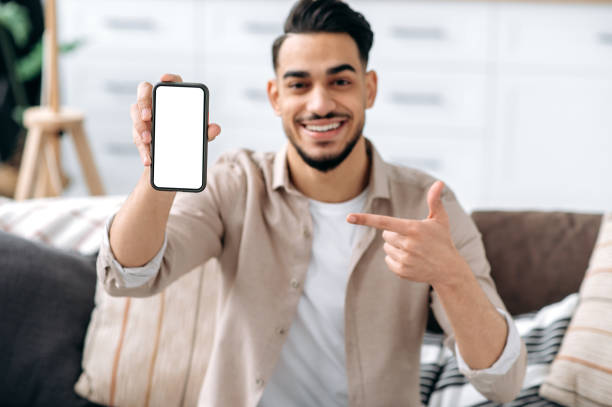 istock Phone screen mockup. Defocused smiling mixed race guy, showing smart phone with empty white mock-up screen, copy-space for advertising or presentation, sitting on a sofa in a living room interior 1418863864