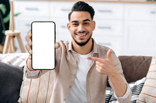 Phone screen mockup. Friendly smiling indian or arabian guy, showing smart phone with empty white mock-up screen, copy-space for advertising or presentation, sits on a sofa in a living room interior