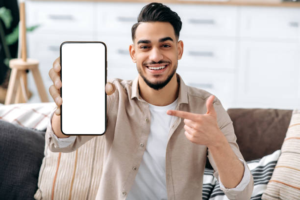 Phone screen mockup. Friendly smiling indian or arabian guy, showing smart phone with empty white mock-up screen, copy-space for advertising or presentation, sits on a sofa in a living room interior stock photo