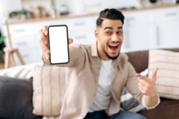 istock Defocused excited arabian or indian young man, holding smart phone with empty white mock-up screen in hand, copy-space for advertising or presentation, sitting on a sofa in a living room interior 1418863895
