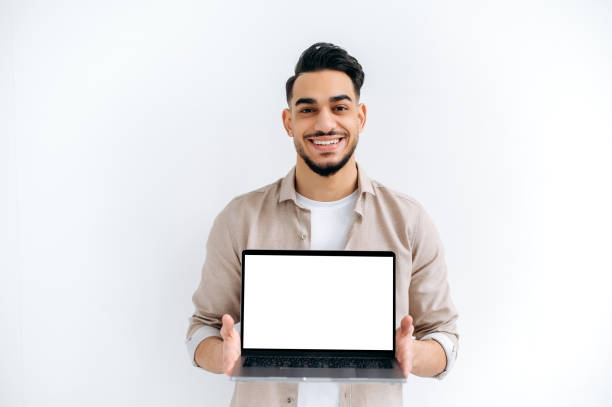 istock Joyful handsome Indian or Arabian young man, student or freelancer, stands on white isolated background, holding open laptop with blank white mockup screen for presentation, looking at camera, smiling 1418863922